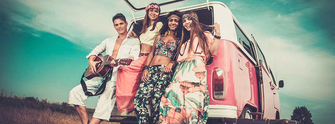 Un grupo de chicos y chicas vestidos con indumentaria hippie se apoyan en el maletero abierto de una furgoneta Wolskvagen rosa. Están sonrientes y el chico sujeta una guitarra. A su lado se encuentran varias maletas.
