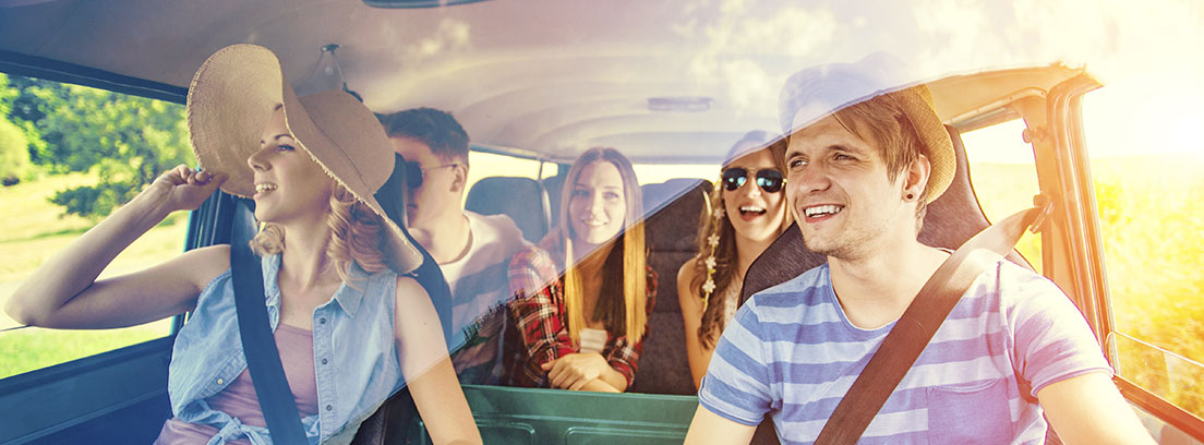 un grupo compuesto por dos chicos y chicas jóvenes sonríen dentro de una furgoneta. Van vestidos con ropa de verano y una de las chicas con una pamela. Miran por una de las ventanillas laterales del vehículo. 