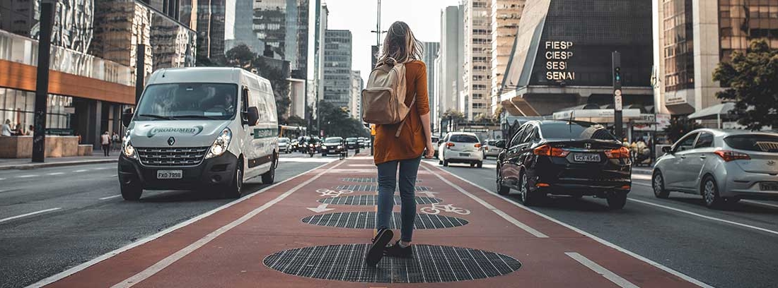 Mujer con mochila en medio de una ciudad y entre dos calles con coches circulando