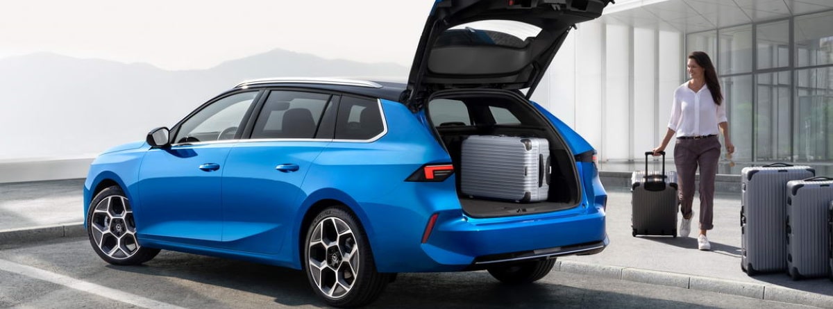 coche azul marca Opel Astra Sports Tourer 2022 con el maletero abierto y una mujer con unas maletas