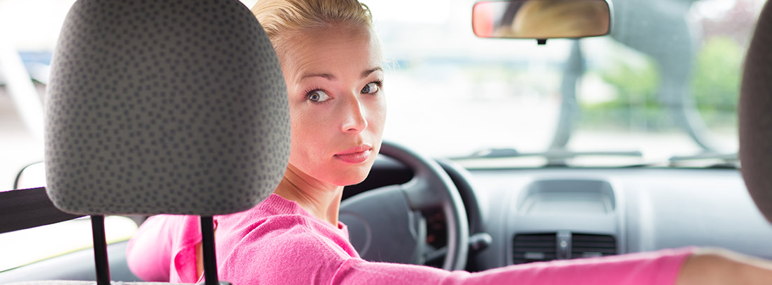 Mujer al volante apoya brazo en asiento copiloto y mira hacia atrás.