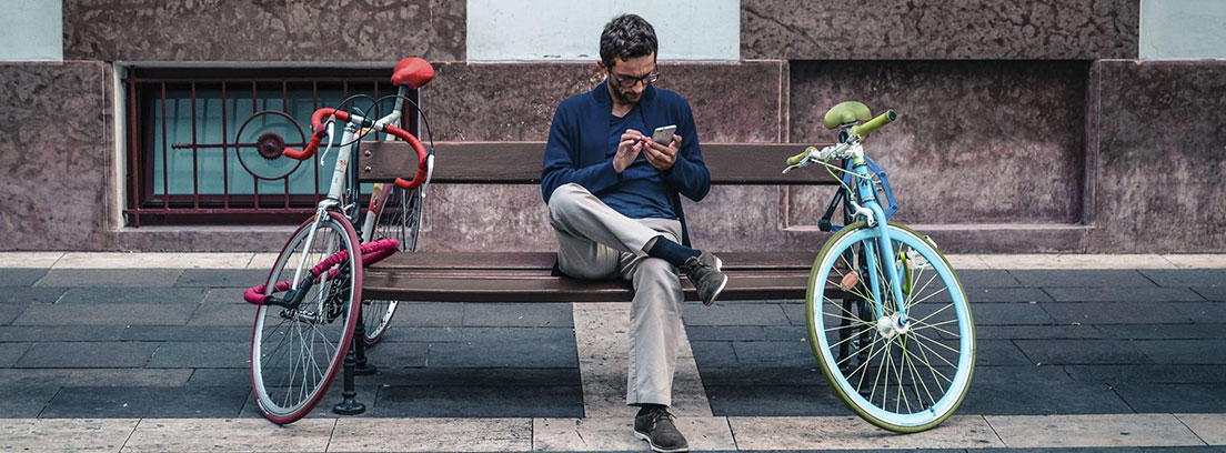 Hombre con gafas mira móvil sentado en banco con dos bicicletas.
