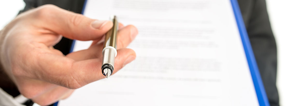 Cláusulas del contrato de compraventa Mano sostiene un bolígrafo hacia delante y unas hojas escritas con fondo blanco.