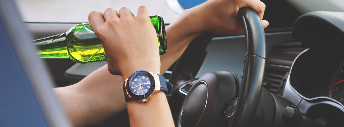 Vista parcial de un hombre bebiendo un botellín de cerveza dentro de un coche con una mano en el volante